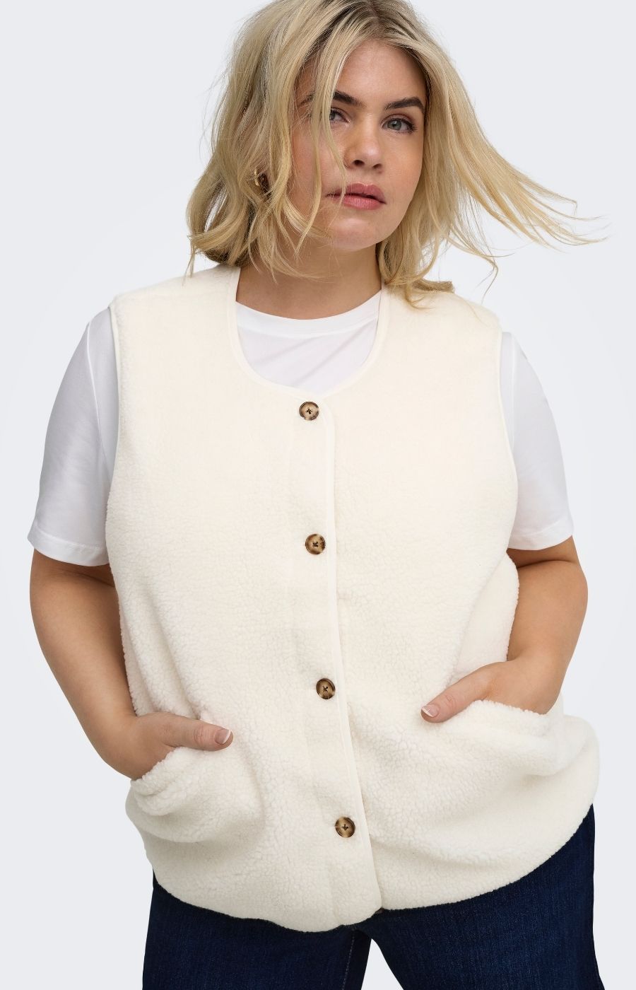 A woman wearing the Only Carmakoma Baby Gilet in Cream, a cream sherpa fleece button-front vest over a white t-shirt and dark blue jeans, photographed against a light gray studio background.