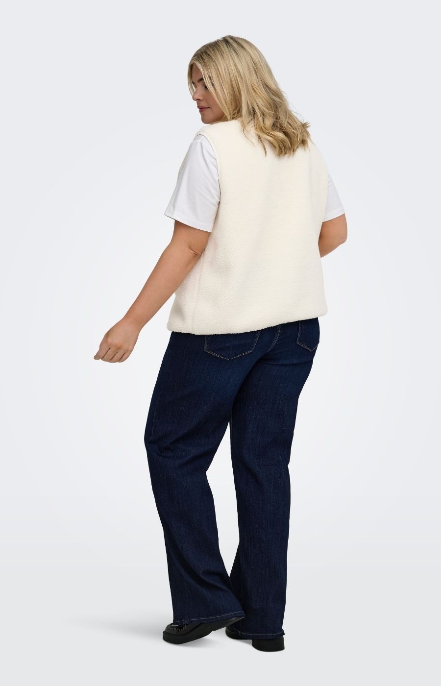 A woman shown from the back wearing the Only Carmakoma Baby Gilet in Cream,  a cream knit sleeveless vest over a white short-sleeve t-shirt and dark blue jeans, standing against a light gray studio background.