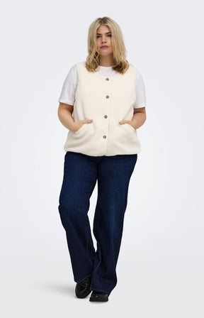 A woman modeling the Only Carmakoma Baby Gilet in Cream, a cream-colored sleeveless button-up vest over a white t-shirt and dark blue jeans, standing against a light gray studio background.he Only Carmakoma Baby Gilet in Cream