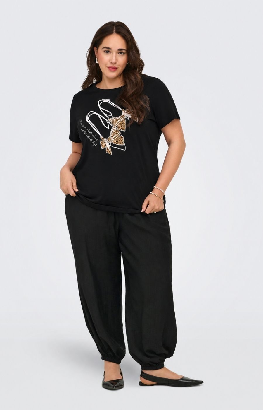 A woman modeling the Only Carmakoma Beau T-Shirt in Black, a black short-sleeve t-shirt with a s shoe motiff, with rhinestone and leopard print detailing, paired with black wide-leg pants and black flats, against a light gray studio background. This image was created using AI.