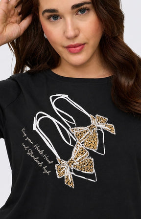A close-up of a woman wearing the Only Carmakoma Beau T-Shirt in Black, a black graphic t-shirt. The shirt features a white line-art graphic of high heels with leopard-print bows, with rhinestone detailing and white script text on the front. This image was created using AI.