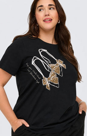 A woman wearing the Only Carmakoma Beau T-Shirt in Black, a black short-sleeve t-shirt with a graphic of two high-heeled shoes in leopard print and white outlines, posed against a light gray background. This image was created using AI.