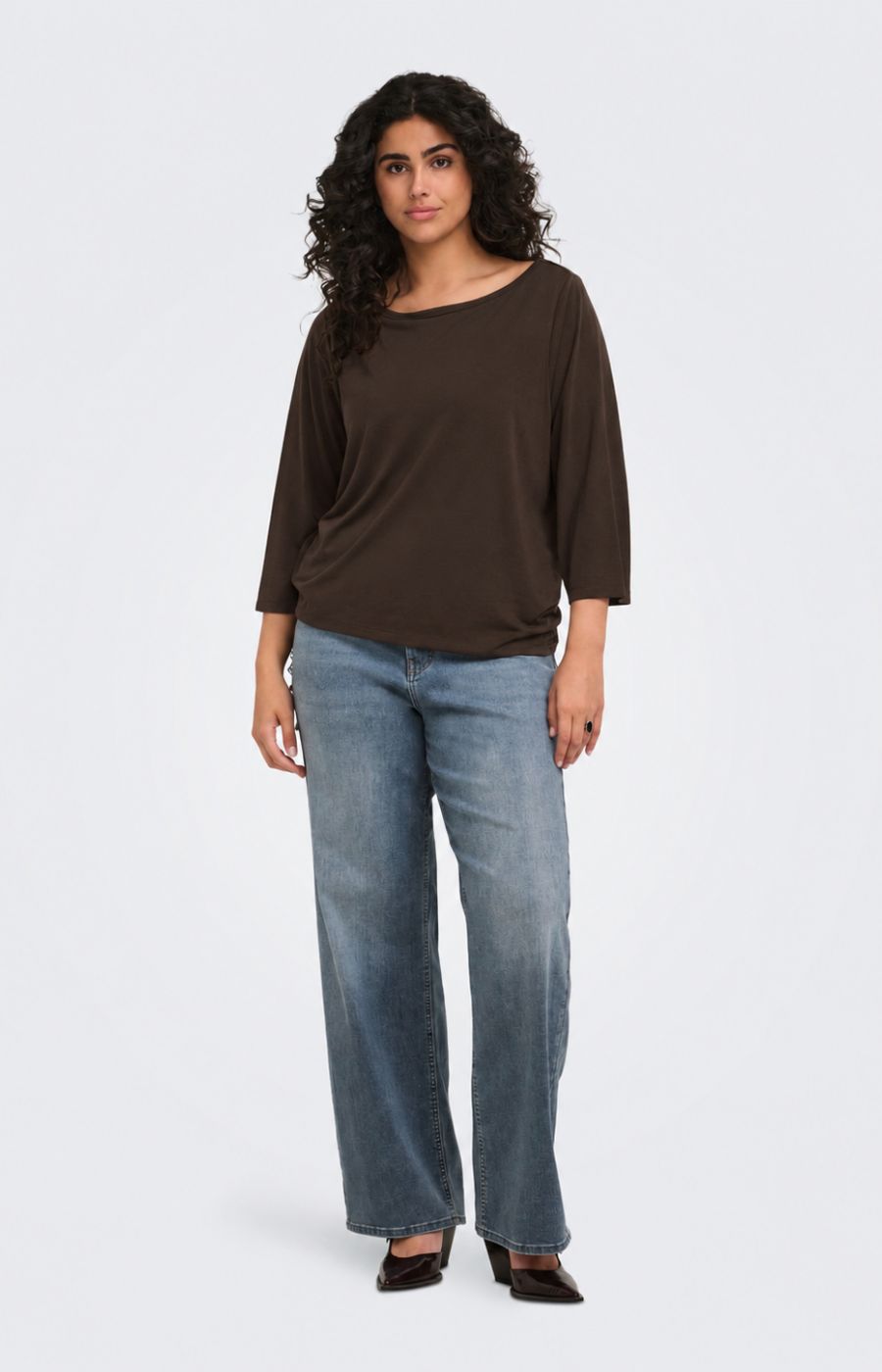 A woman modeling the Only Carmakoma Carlinne Top in Coffee Bean, a dark brown boat-neck top with three-quarter sleeves and light-wash blue jeans, standing against a light gray studio background.