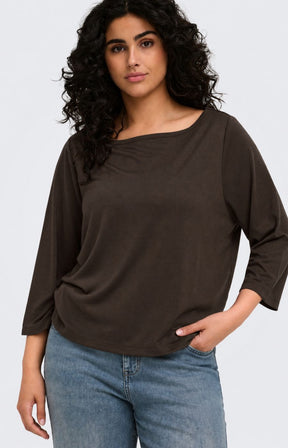 A woman wearing the Only Carmakoma Carlinne Top in Coffee Bean, a dark brown, boat-neck, three-quarter sleeve top and blue jeans, photographed against a light gray studio background.
