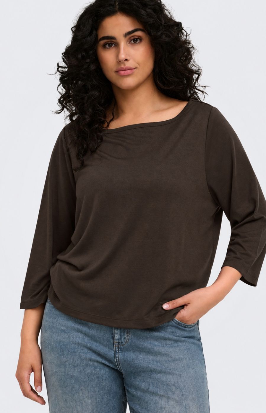 A woman wearing the Only Carmakoma Carlinne Top in Coffee Bean, a dark brown, boat-neck, three-quarter sleeve top and blue jeans, photographed against a light gray studio background.
