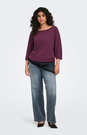 A woman modeling the Only Carmakoma Carlinne Top in Fig,  a loose-fitting, three-quarter sleeve boat-neck top in deep plum purple, paired with wide-leg blue jeans and black shoes, against a plain light gray studio background.