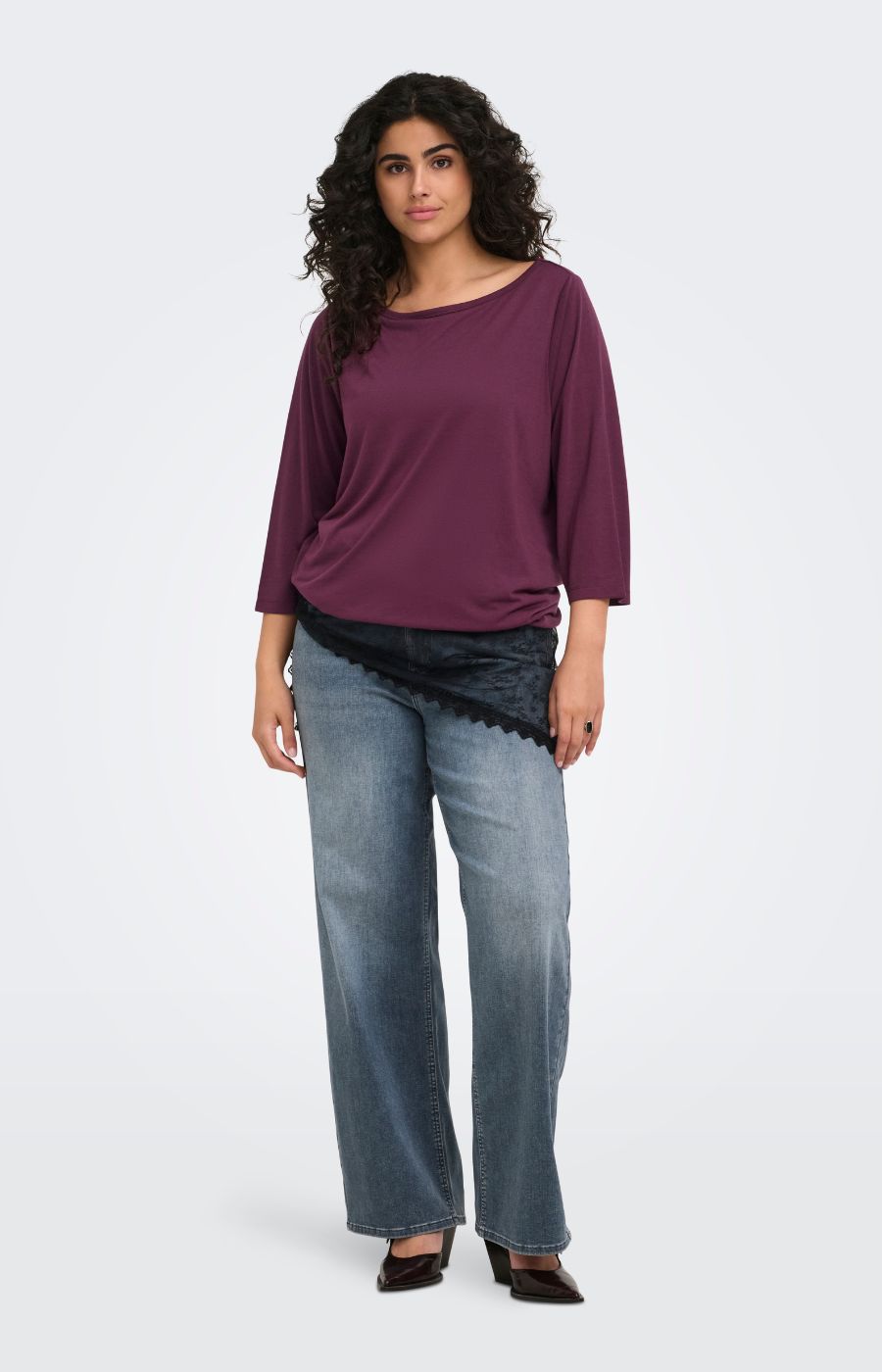 A woman modeling the Only Carmakoma Carlinne Top in Fig,  a loose-fitting, three-quarter sleeve boat-neck top in deep plum purple, paired with wide-leg blue jeans and black shoes, against a plain light gray studio background.
