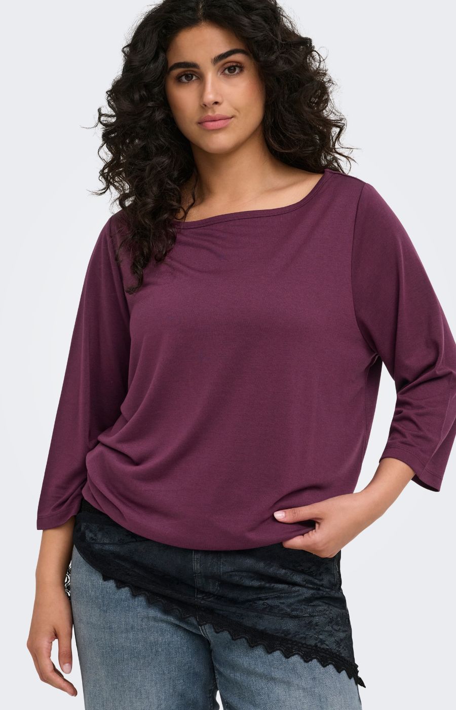 A woman wearing the Only Carmakoma Carlinne Top in Fig, a loose-fitting, three-quarter sleeve top in a deep plum purple color, paired with blue jeans and a black lace-trimmed sash, against a plain light gray background.