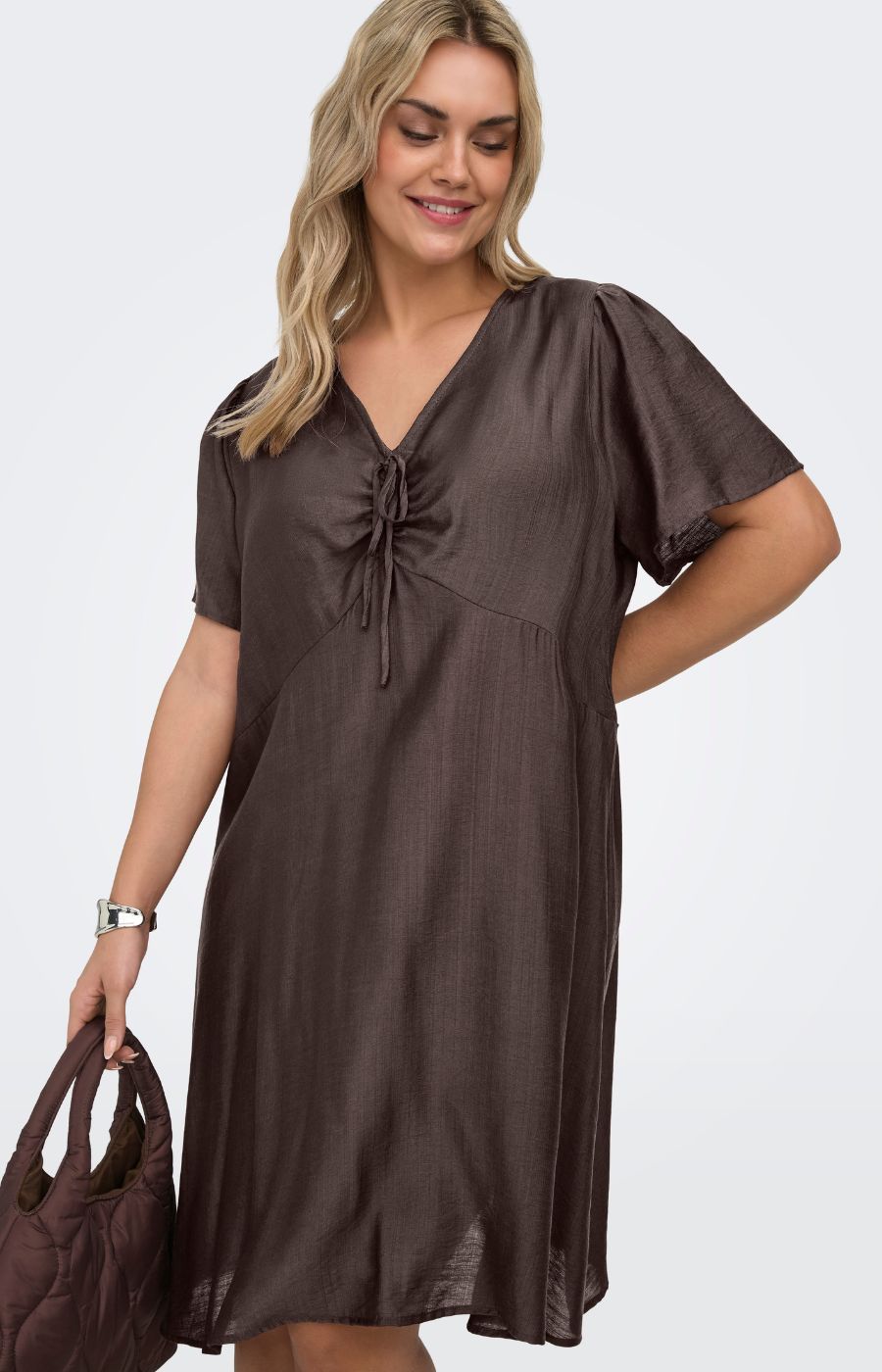 Woman wearing the Only Carmakoma Cecilia Dress in Brown, a brown dress holding a matching handbag on a white background