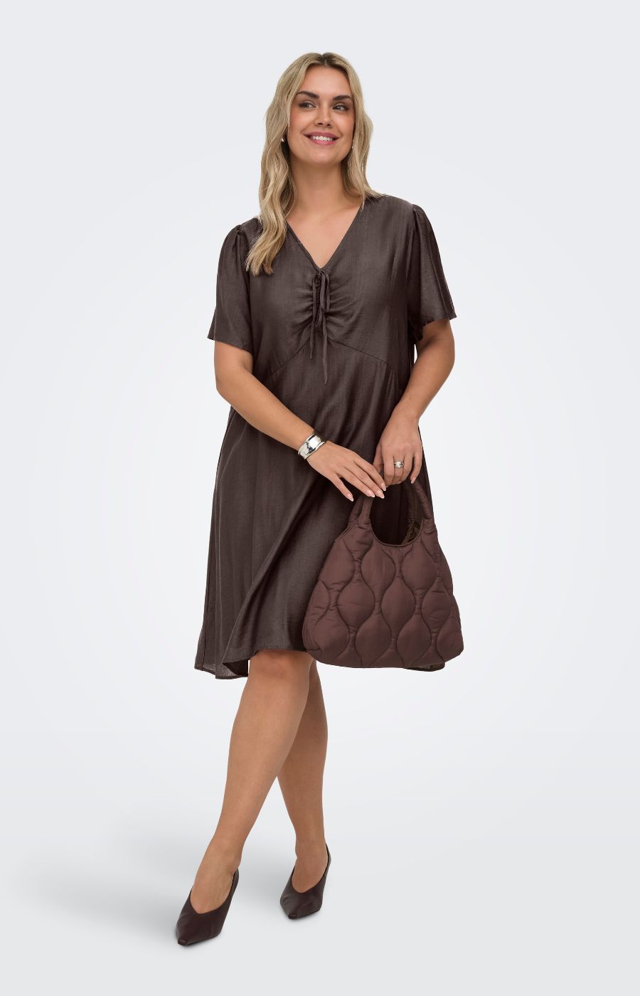 Woman wearing the Only Carmakoma Cecilia Dress in Brown, a brown dress holding a matching handbag on a white background