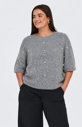 A woman wearing the Only Carmakoma Dolly Top in Grey a grey knit sweater with pearl-like bead embellishments and black trousers, standing against a light gray studio background. This image was created using AI.