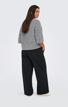 A woman wearing the Only Carmakoma Dolly Top in Grey a grey knit sweater with pearl-like bead embellishments and black trousers, standing against a light gray studio background. This image was created using AI.