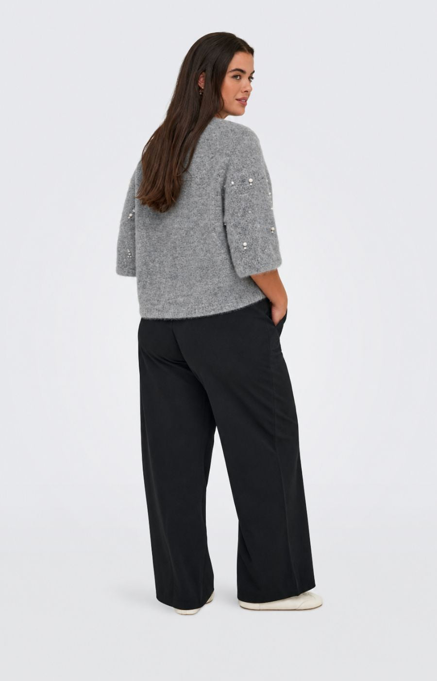 A woman wearing the Only Carmakoma Dolly Top in Grey a grey knit sweater with pearl-like bead embellishments and black trousers, standing against a light gray studio background. This image was created using AI.