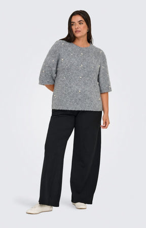 A woman wearing the Only Carmakoma Dolly Top in Grey, a grey knit sweater with pearl-like bead embellishments and black trousers, standing against a light gray studio background. This image was created using AI.