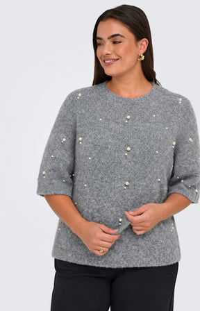A woman wearing the Only Carmakoma Dolly Top in Grey, a grey knit sweater with pearl-like bead embellishments and black trousers, standing against a light gray studio background. This image was created using AI.