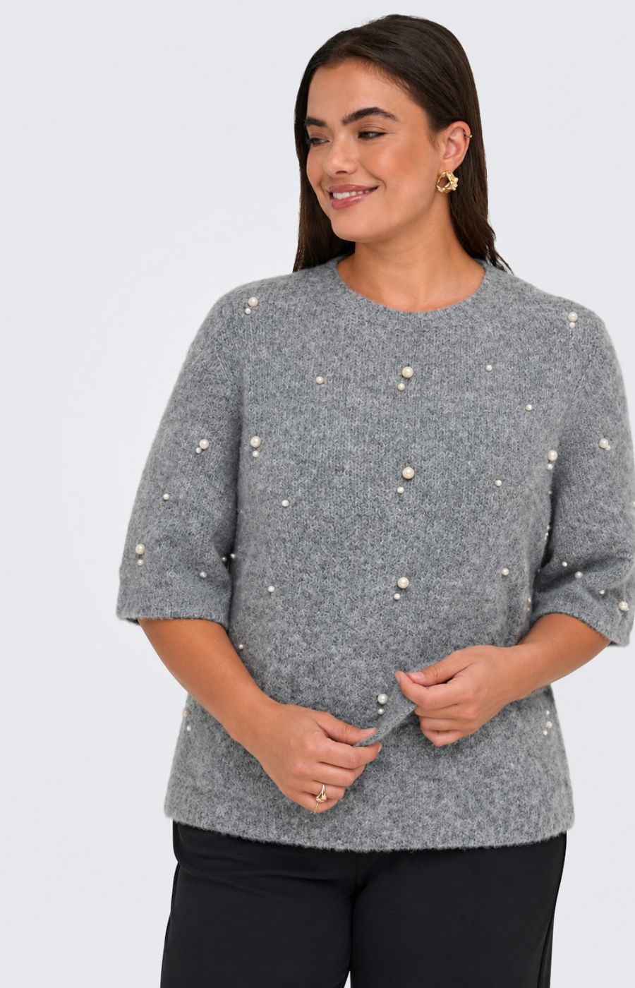 A woman wearing the Only Carmakoma Dolly Top in Grey, a grey knit sweater with pearl-like bead embellishments and black trousers, standing against a light gray studio background. This image was created using AI.