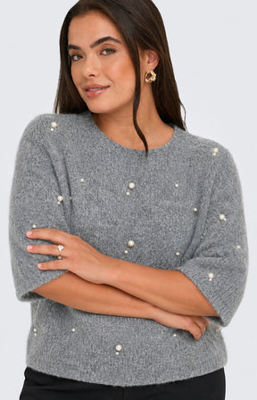 A woman wearing the Only Carmakoma Dolly Top in Grey, a grey knit sweater with pearl-like bead embellishments and black trousers, standing against a light gray studio background. This image was created using AI.