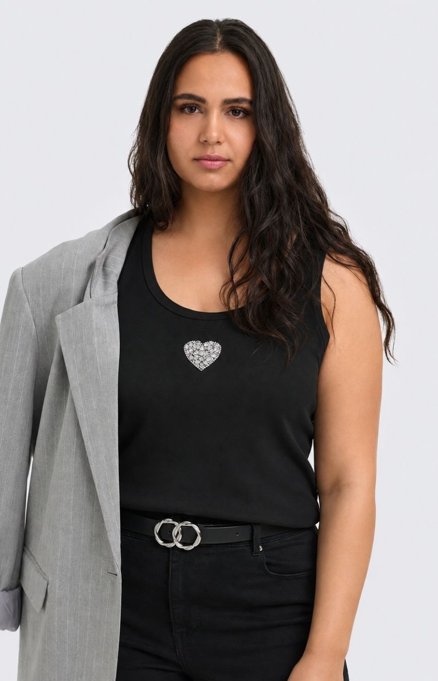 A woman wearing the Only Carmakoma Iris Vest Top in Black, a black sleeveless tank top with a silver heart-shaped embellishment on the chest, styled with a gray blazer and black pants against a light gray background. This image was created using AI.