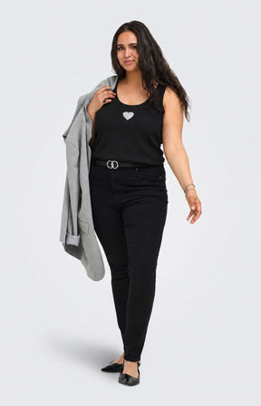 A woman modeling the Only Carmakoma Iris Vest Top in Black, a black sleeveless top with a silver heart emblem, black skinny pants, a black belt with silver rings, and black flats, while holding a light gray blazer against a plain light gray background. This image was created using AI.