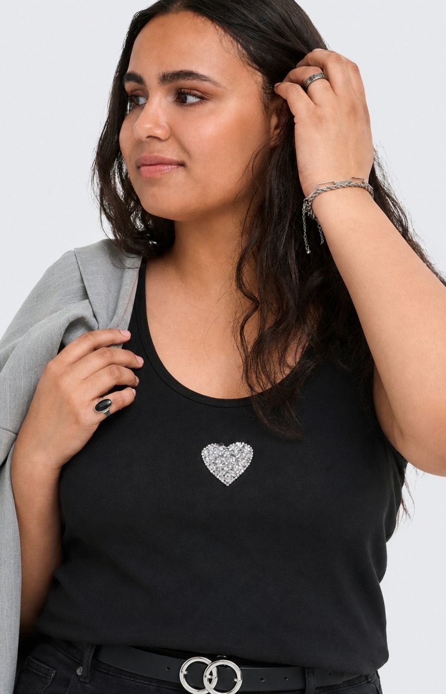 A woman wearing the Only Carmakoma Iris Vest Top in Black, a black tank top with a sparkling silver heart-shaped brooch on the chest, styled with a gray cardigan and black pants against a light gray background. This image was created using AI.