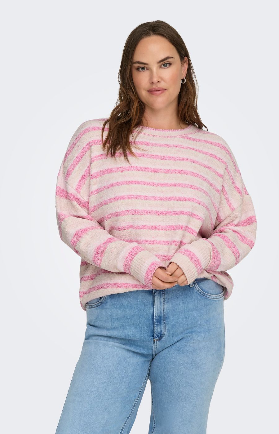 Woman wearing the Only Carmakoma Johanna Knit in Pink, a pink and white striped sweater with long sleeves and a round neckline, styled with blue jeans on a light gray background