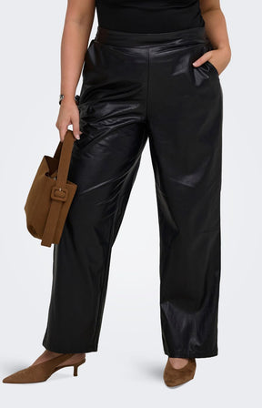 A model wearing the Only Carmakoma Kari Trousers in Black, black straight leg faux leather pants, styled with a black top, brown suede shoes, and a brown leather tote bag, set against a plain light grey studio background.