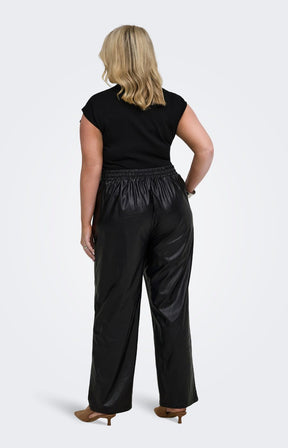 A woman viewed from the back wearing the Only Carmakoma Kari Trousers in Black, black straight leg pants with a shiny finish, a black short-sleeve top, and brown heels, against a light grey studio background.
