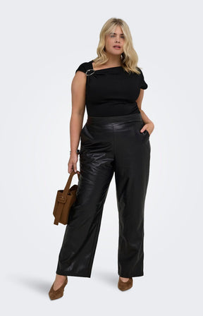 A woman modelling the Only Carmakoma Kari Trousers in Black, black faux-leather straight-leg trousers, a black off-the-shoulder short-sleeve top and holding a brown handbag against a light grey studio background.