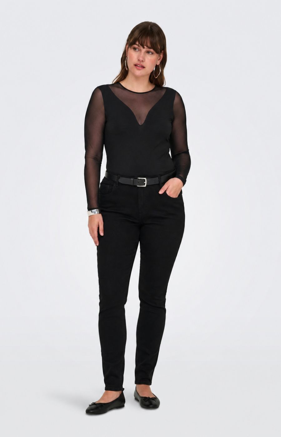 A woman modeling the Only Carmakoma Love Top in Black, a black sheer long-sleeve top with a V-neckline, paired with black skinny jeans, a black belt, and black flats, against a light gray studio background.