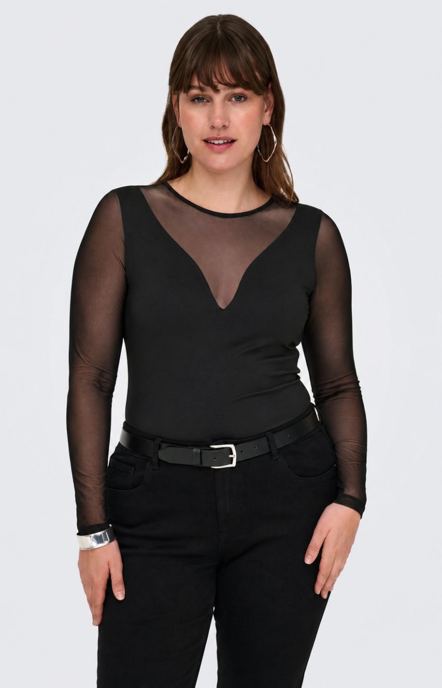 A woman modelling the Only Carmakoma Love Top in Black, a black long-sleeve bodysuit with sheer mesh sleeves and a sheer V-neckline, paired with black pants and a black belt, against a light gray studio background.