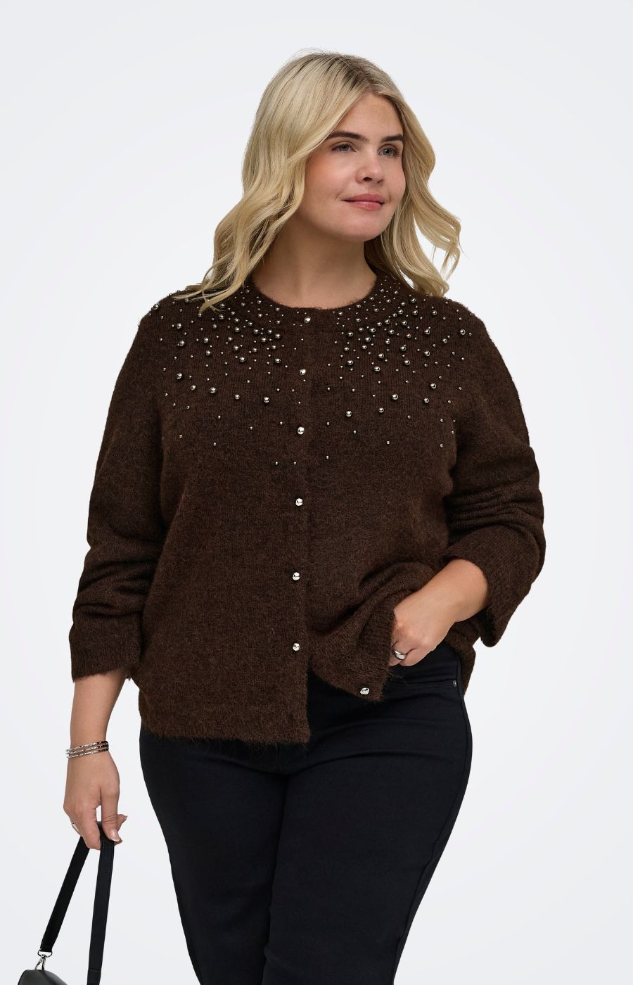 A woman modeling  the Only Carmakoma Miggie Cardigan in Coffee Bean, a dark brown knit cardigan sweater with silver rhinestone embellishments across the chest, paired with black pants against a plain white studio background.