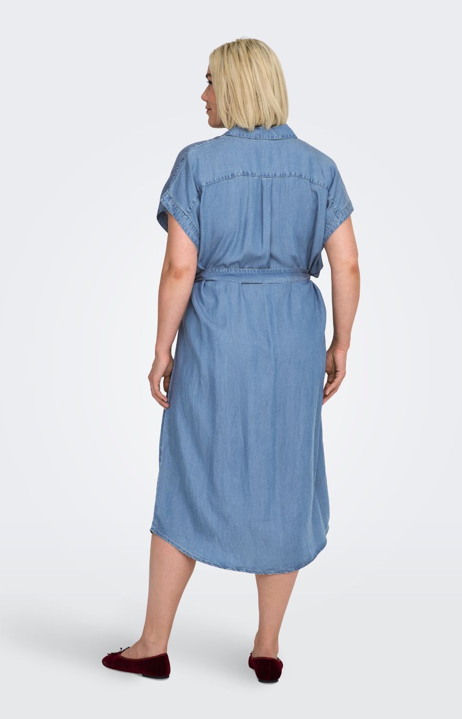 Woman wearing the Only Carmakoma Pema Denim Dress, a blue midi dress with a removable tie belt on a white background
