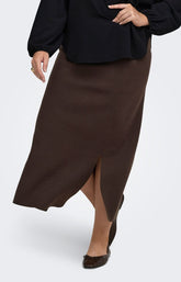 A woman wearing the Only Carmakoma Ribi Skirt in Brown, a dark brown maxi skirt with a front slit, paired with a black long-sleeve blouse and dark brown pointed flats, shown against a light gray studio background.