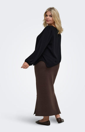 A woman modeling the Only Carmakoma Ribi Skirt in Brown, a knitted brown maxi skirt and a black long-sleeve top, shown from a side-back angle against a light gray studio background.