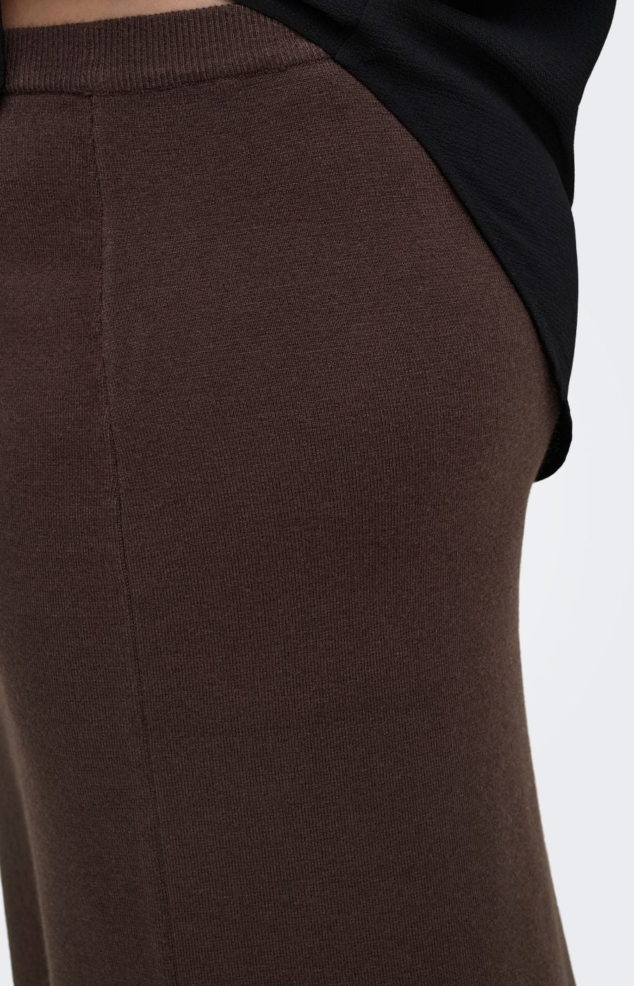 Close-up of a person wearing the Only Carmakoma Ribi Skirt in Brown, a dark brown ribbed knit skirt  with a black top visible at the waist, against a plain light background. 