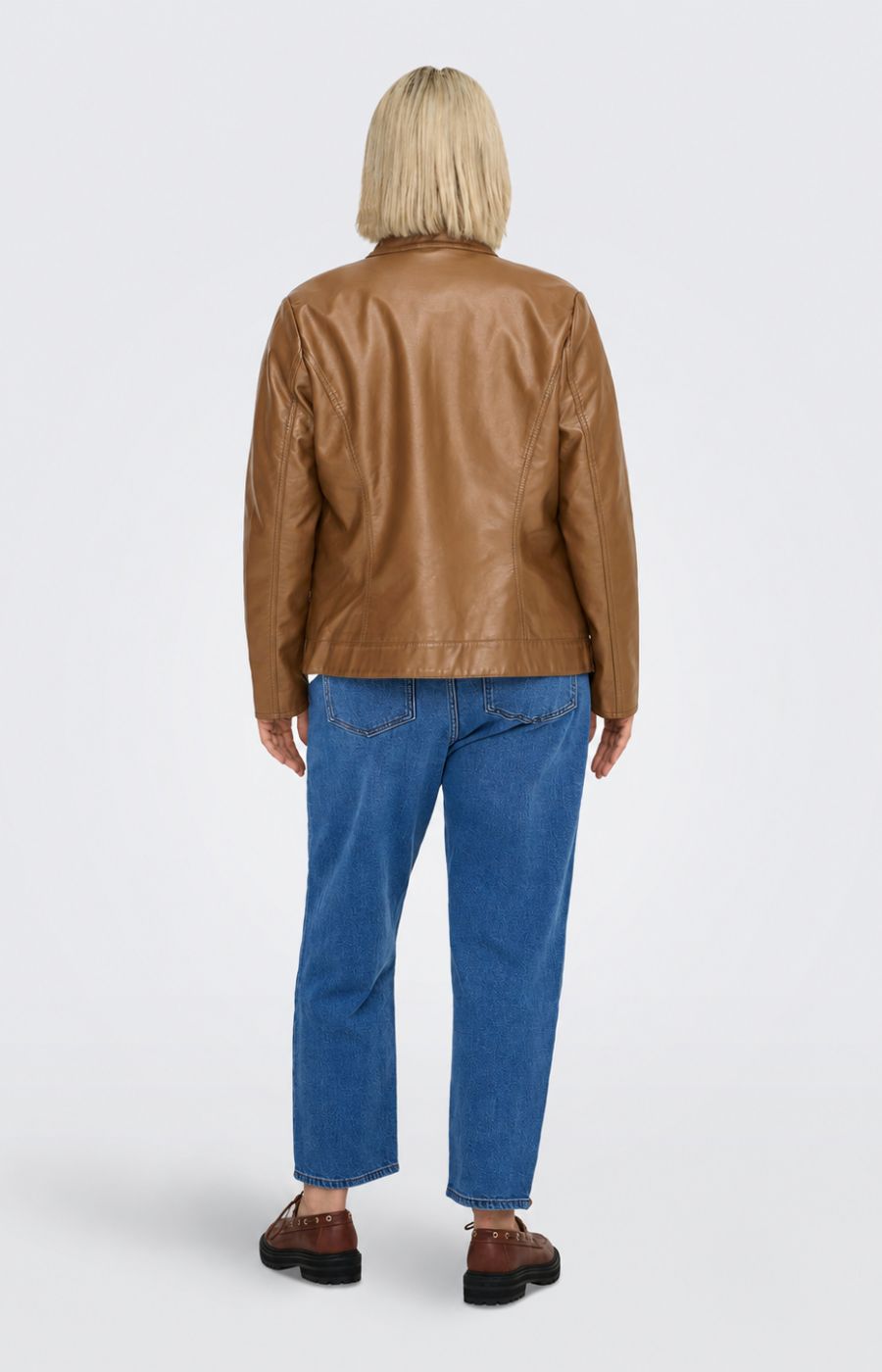 A woman modeling the Only Carmakoma Robber Faux Leather Jacket in Cognac, a cropped tan faux-leather jacket over a white top and blue jeans, standing against a light gray studio background. This image was created using AI.