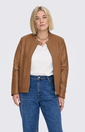 A woman modeling the Only Carmakoma Robber Faux Leather Jacket in Cognac, a cropped tan faux-leather jacket over a white top and blue jeans, standing against a light gray studio background. This image was created using AI.