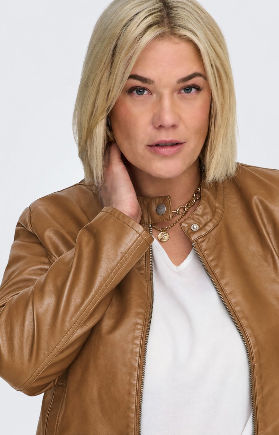 A woman modeling the Only Carmakoma Robber Faux Leather Jacket in Cognac, a cropped tan faux-leather jacket over a white top and blue jeans, standing against a light gray studio background. This image was created using AI.