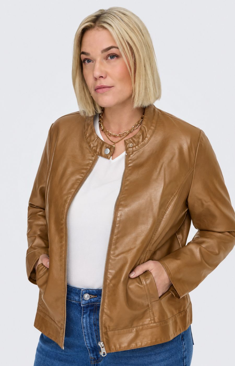 A woman modeling the Only Carmakoma Robber Faux Leather Jacket in Cognac, a cropped tan faux-leather jacket over a white top and blue jeans, standing against a light gray studio background. This image was created using AI.