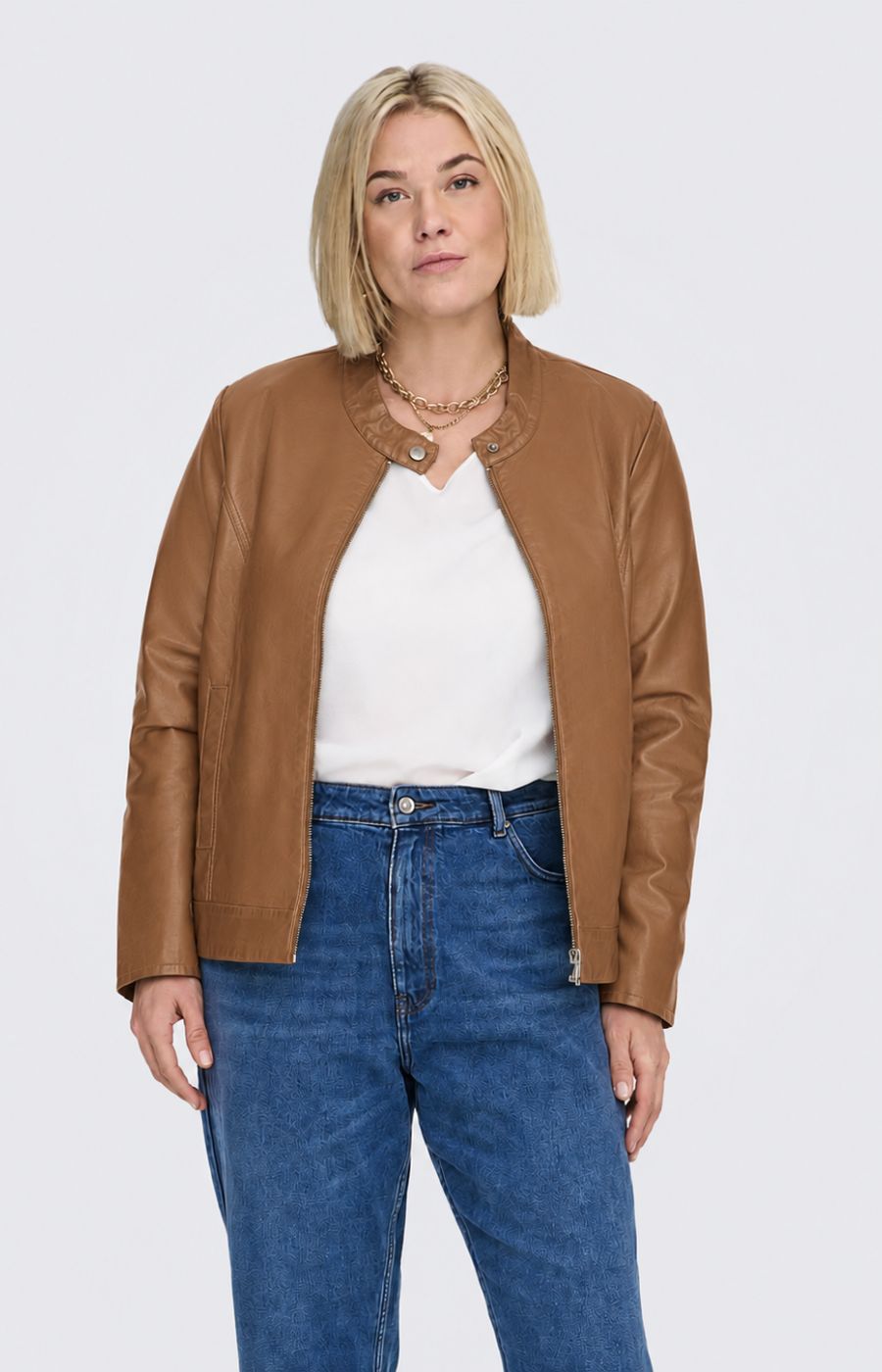 A woman modeling the Only Carmakoma Robber Faux Leather Jacket in Cognac, a cropped tan faux-leather jacket over a white top and blue jeans, standing against a light gray studio background. This image was created using AI.