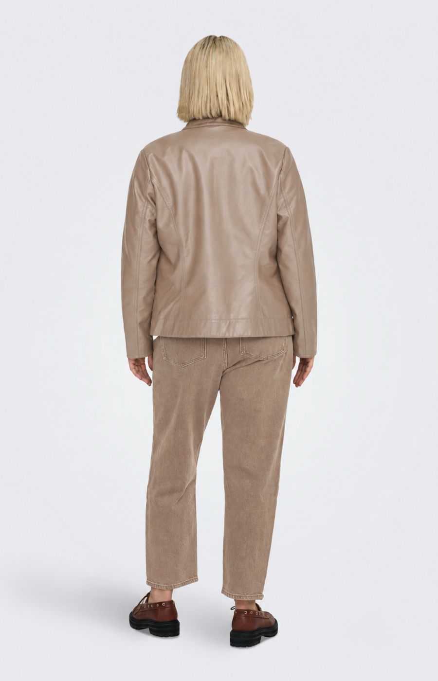 A woman modeling the Only Carmakoma Robber Faux Leather Jacket in Walnut, a light taupe faux-leather jacket with a zippered front, buttoned collar, and side pockets, paired with a white top and matching taupe pants against a light gray background. This image has be created using AI.