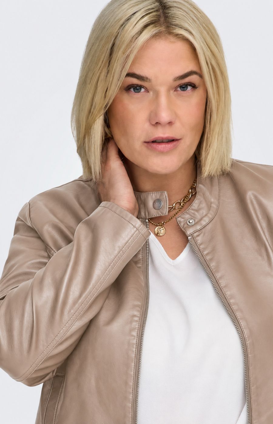 A woman modeling the Only Carmakoma Robber Faux Leather Jacket in Walnut, a light taupe faux-leather jacket with a zippered front, buttoned collar, and side pockets, paired with a white top and matching taupe pants against a light gray background. This image has be created using AI.