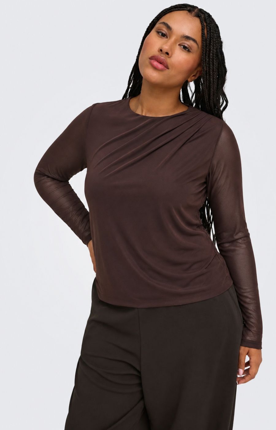 A woman modeling the Only Carmakoma Sara Top in Chocolate, a long-sleeve, round neck top with draping on the shoulder, in a dark brown color, paired with dark brown pants, against a light gray studio background. This image was created using AI.