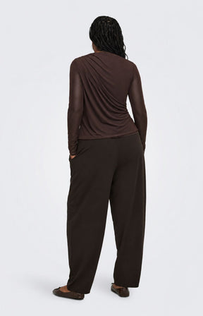 A model viewed from the back wearing the Only Carmakoma Sara Top in Chocolate, a dark brown draped long-sleeve top and matching dark brown wide-leg pants against a light gray studio background.