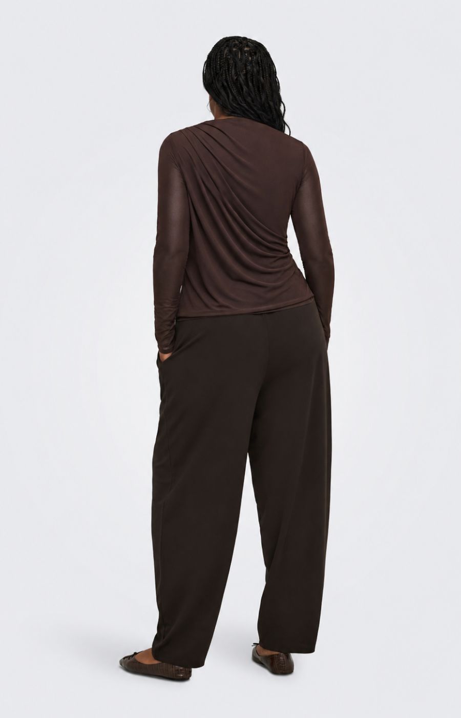 A model viewed from the back wearing the Only Carmakoma Sara Top in Chocolate, a dark brown draped long-sleeve top and matching dark brown wide-leg pants against a light gray studio background.