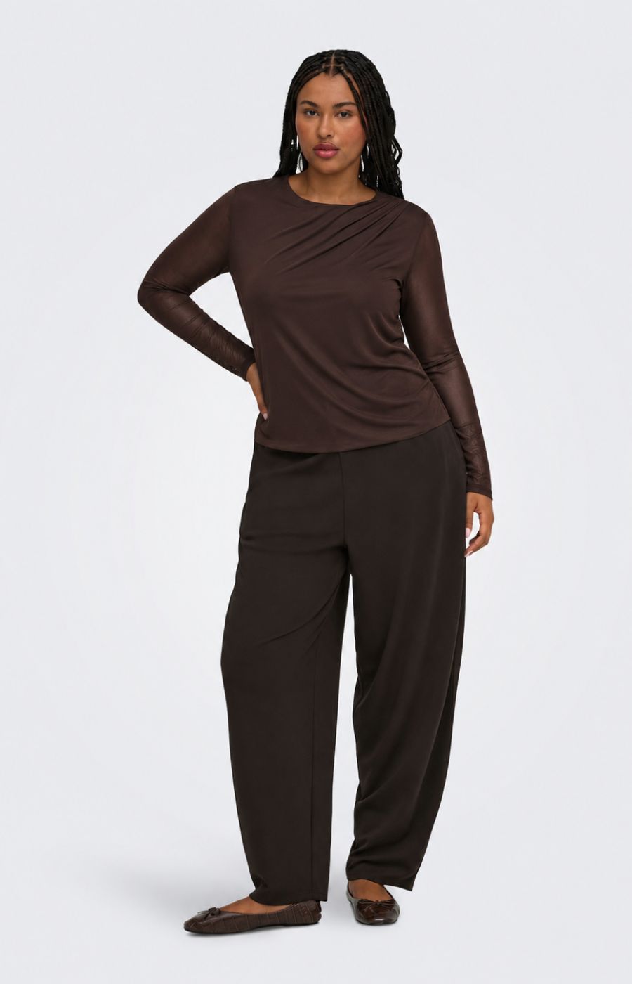 A woman modeling the Only Carmakoma Sara Top in Chocolate, a dark brown long-sleeve top and matching dark brown wide-leg pants against a light gray studio background.