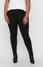 A model wearing the Only Carmakoma Tay Trousers in Black, black skinny leg trousers with a white top and black ankle boots, shown from the chest down against a light gray studio background.