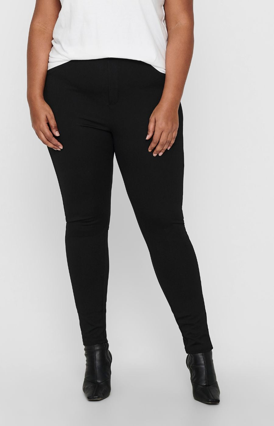 A model wearing the Only Carmakoma Tay Trousers in Black, black skinny leg trousers with a white top and black ankle boots, shown from the chest down against a light gray studio background.