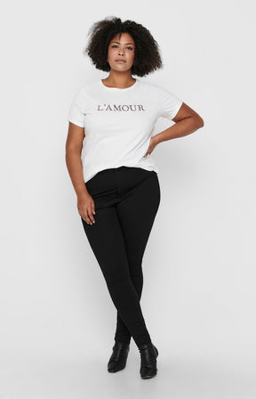 A woman modeling the Only Carmakoma Tay Trousers in Black with a white short-sleeve t-shirt with 'L'AMOUR' printed in black across the chest, black ankle boots, against a light gray studio background.