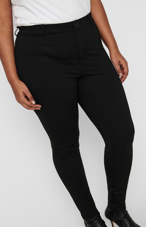 A model wearing the Only Carmakoma Tay Trousers in Black,  black slim-fit skinny pants with a white top and black boots, shown from the waist down against a light gray background.